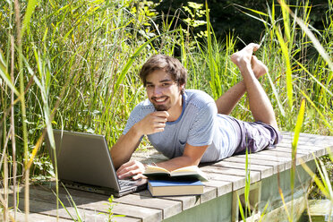 Porträt eines lächelnden jungen Mannes, der mit Büchern und Laptop auf einer Holzpromenade liegt - WWF003838