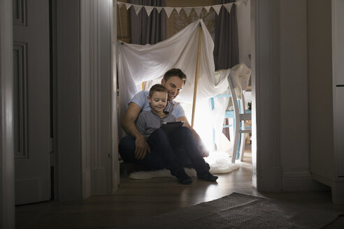 Father and son sitting in self-made tent at home in the evening using digital tablet - PDF000685