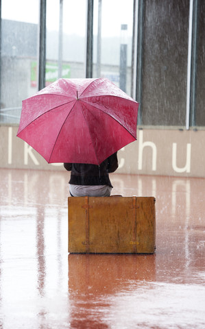 Österreich, Thalgau, junges Mädchen mit rotem Regenschirm auf ihrem Koffer sitzend im Regen, lizenzfreies Stockfoto