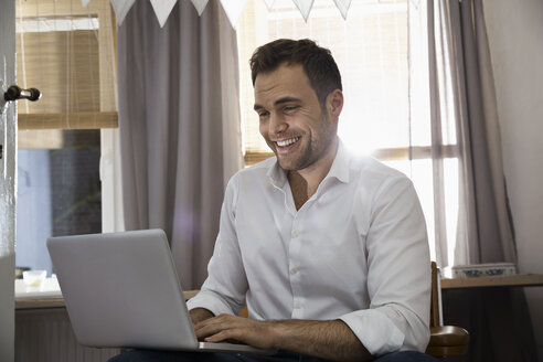 Lachender Mann bei der Arbeit mit Laptop in seinem Heimbüro - PDF000706