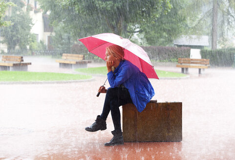 Austria, Thalgau, teenage girl with red umbrella sitting on her suitcase in the rain - WWF003771