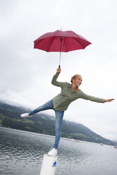 Österreich, Mondsee, junges Mädchen mit rotem Regenschirm balanciert auf einer Stange vor einem See - WWF003781