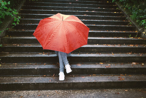 Teenage girl sitting on stairs hiding behind red umbrella - WWF003792