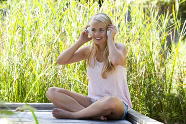 Portrait of smiling young woman with headphones sitting on wooden boardwalk - WWF003844