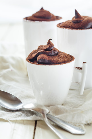 Drei Tassen vegane Mousse au Chocolat, Teelöffel, Stoff auf weißem Holz, lizenzfreies Stockfoto