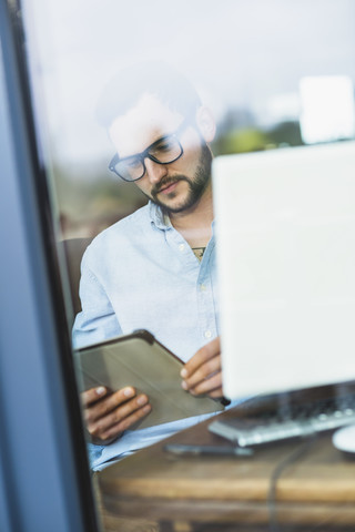 Junger Mann hinter einer Fensterscheibe mit Blick auf ein digitales Tablet, lizenzfreies Stockfoto