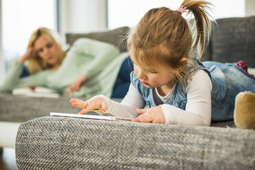 Mädchen benutzt digitales Tablet auf Couch mit Mutter im Hintergrund - UUF003407