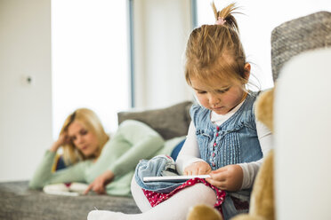 Mädchen benutzt digitales Tablet auf Couch mit Mutter im Hintergrund - UUF003403