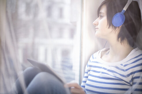 Young woman with headphones and digital tablet looking through window - SEGF000226