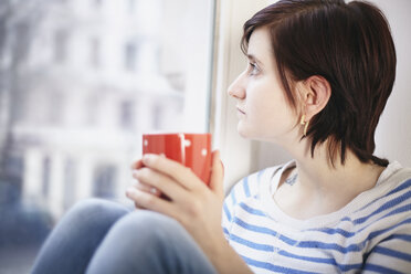 Junge Frau mit einer Tasse Kaffee schaut durch ein Fenster - SEGF000227