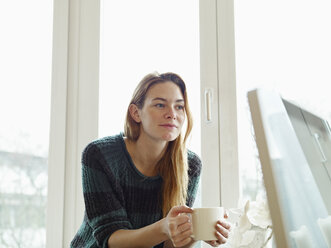 Deutschland, Köln, Junge Frau sitzt am Fenster und hält eine Tasse Kaffee - RHF000550