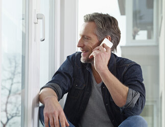 Germany, Cologne, Mature man sitting at window using smart phone - RHF000492