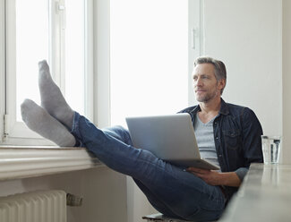 Deutschland, Köln, Älterer Mann am Fenster sitzend mit Laptop, Füße hochgelegt - RHF000511