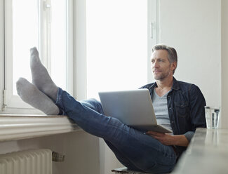 Germany, Cologne, Mature man sitting at window using laptop, feet up - RHF000511