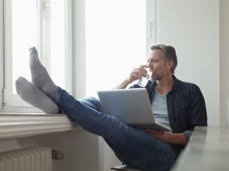 Deutschland, Köln, Älterer Mann am Fenster sitzend mit Laptop, Füße hochgelegt - RHF000512