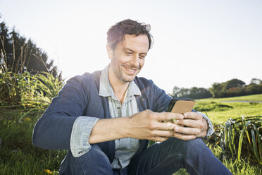 Smiling man sitting on a meadow reading SMS - PDF000802