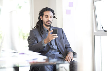 Businessman with dreadlocks sitting in an office looking at smartphone - ZEF003518