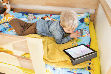 Kleiner Junge liegt auf seinem Bett und schaut auf ein digitales Tablet mit einer Kinderzeichnung - MFF001490