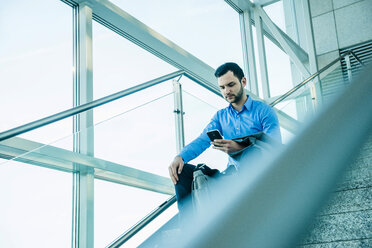 Young businessman sitting on stairs looking on cell phone - UUF003456