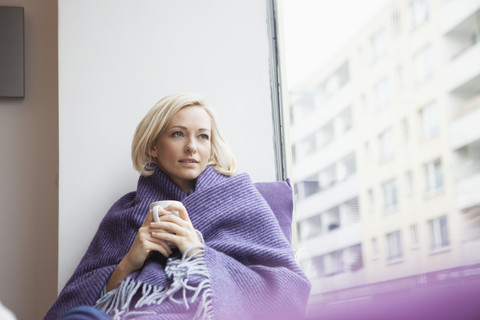 Porträt einer Frau mit Tasse und Wolldecke, die durch ein Fenster schaut, lizenzfreies Stockfoto