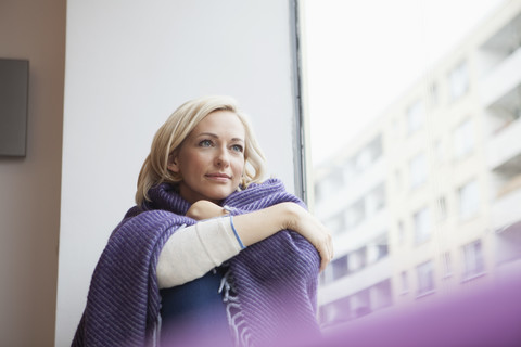 Porträt einer Frau mit Wolldecke, die durch ein Fenster schaut, lizenzfreies Stockfoto