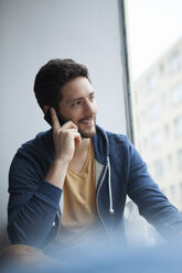 Porträt eines lächelnden Mannes, der mit seinem Smartphone telefoniert und durch ein Fenster schaut - RBF002483