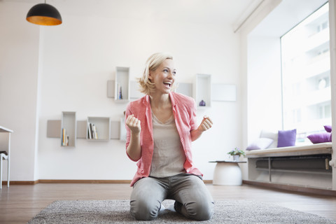 Jubelnde Frau auf Teppich im Wohnzimmer sitzend, lizenzfreies Stockfoto