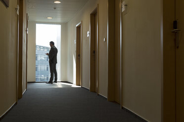 Business man standing in corridor checking mobile phone - WESTF020847