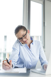 Businesswoman sitting at desk taking notes on the phone - WESTF020894