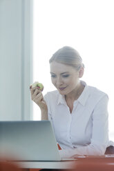 Businesswoman sitting at desk eating apple - WESTF020978