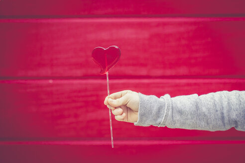 Close-up of boy holding heart-shaped lollipop in front of red wall - SARF001433
