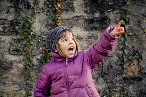 Excited girl wearing woolly hat and pink winter jacket pointing at something - LVF002945