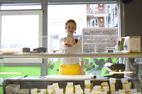 Lächelnde junge Frau in einem Naturkostladen, die ein Stück Käse überreicht - SGF001387