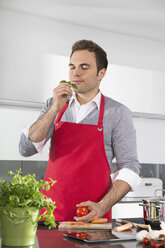 Portrait of man with red apron standing in kitchen smelling basil - PDF000832