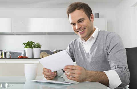 Lächelnder Mann, der in der Küche sitzt und den Briefumschlag betrachtet, lizenzfreies Stockfoto