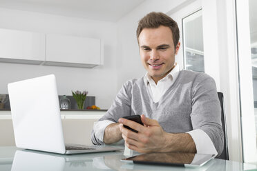 Smiling man sitting in kitchen with laptop using smartphone - PDF000855