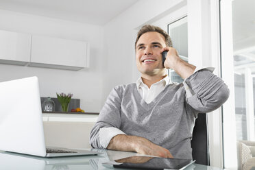 Lächelnder Mann sitzt in der Küche mit Laptop und digitalem Tablet und telefoniert mit einem Smartphone - PDF000856