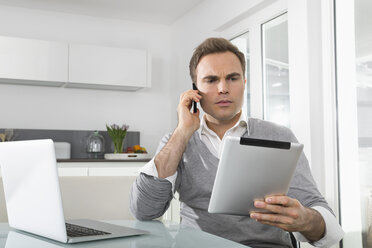 Man sitting in kitchen with laptop using digital tablet and smartphone - PDF000857