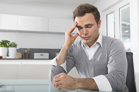 Man with closed eyes sitting in kitchen with hand on his face stock photo