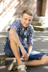 Lächelnder Jugendlicher auf Skateboard sitzend - DRF001480