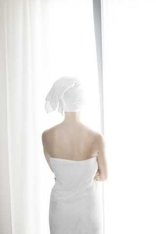 In ein weißes Handtuch gewickelte Frau vor einem weißen Vorhang stehend, lizenzfreies Stockfoto