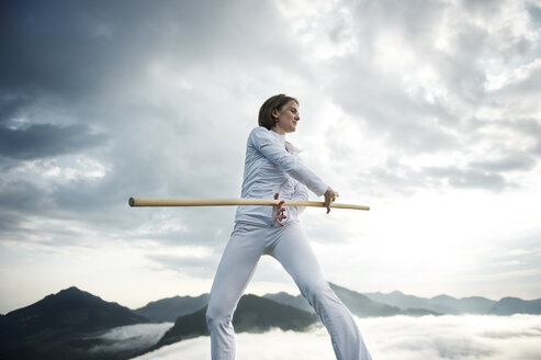 Austria, Kranzhorn, Mid adult woman exercising stick fighting on mountain top - MAOF000033