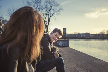 Germany, Berlin, teenage couple sitting face to face at lakeshore in the late afternoon - MMFF000531