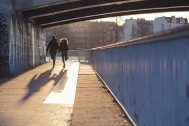 Deutschland, Berlin, jugendliches Paar läuft unter einer Brücke im abendlichen Sonnenlicht - MMFF000528