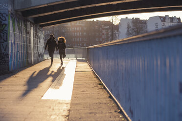 Germany, Berlin, teenage couple running under a bridge at evening sunlight - MMFF000528