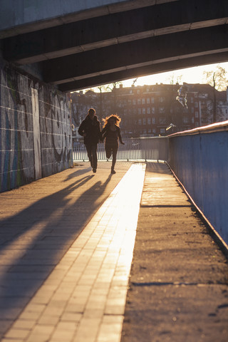 Deutschland, Berlin, jugendliches Paar läuft unter einer Brücke im abendlichen Sonnenlicht, lizenzfreies Stockfoto