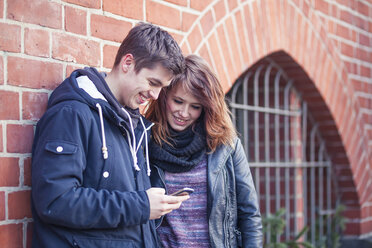 Ein junges Paar steht vor einem Backsteinhaus und beobachtet sein Smartphone - MMFF000509