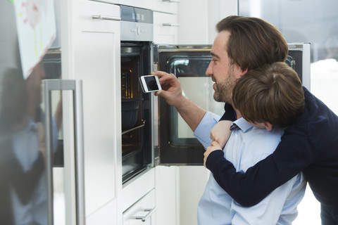 Vater und Sohn fotografieren Kuchen im Ofen mit Smartphone, lizenzfreies Stockfoto
