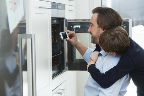 Father and son photographing cake in the oven with smartphone - JTLF000077