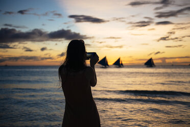 Philippinen, Boracay, Frau beim Fotografieren eines Sonnenuntergangs mit Segelbooten - GEMF000123